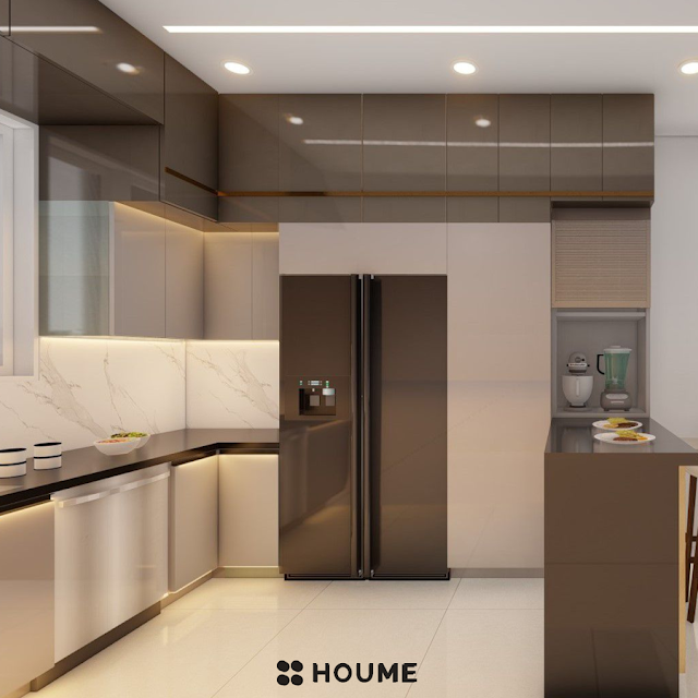 Modern kitchen with a large waterfall island breakfast bar in white quartz, stainless steel appliances, white cabinets with black pulls, and a window with a black frame overlooking a green backyard. Text overlay: Houme India - Kitchen Design Ideas. B