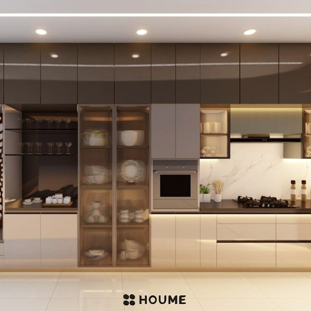 Modern kitchen with a large waterfall island breakfast bar in white quartz, stainless steel appliances, white cabinets with black pulls, and a window with a black frame overlooking a green backyard. Text overlay: Houme India - Kitchen Design Ideas. C