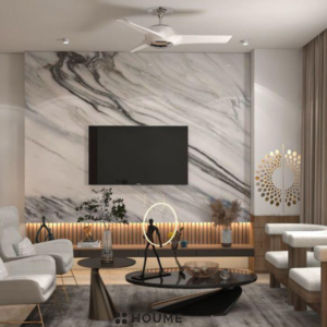 Modern living room with a large flat screen TV mounted on a white wall above a wooden media console. A light gray fabric sofa sits on a woven jute rug in the center of the space. Text overlay: Houme India - Interior Design Ideas. B