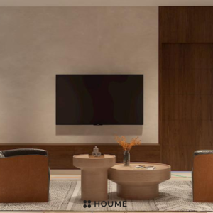 Living room with a modern gray couch, two brown accent chairs, and a round wooden coffee table with black metal legs. A woven jute rug sits underneath the furniture, and a large plant adds a touch of nature to the space. Text overlay: Houme India - Interior Design Ideas. C