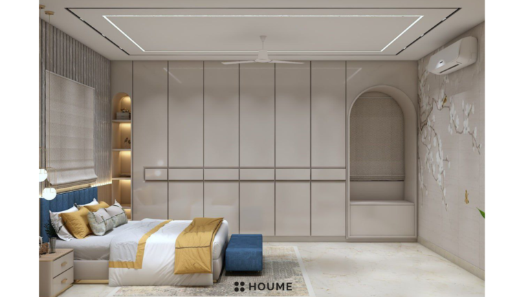 Simple and clean master bedroom interior featuring a platform bed with a light wood frame, white bedding, and a round wooden nightstand with a woven basket underneath. A woven jute rug in a light natural color covers the floor. The walls are painted a light gray color. Text overlay: Houme India - Interior Design Ideas. C