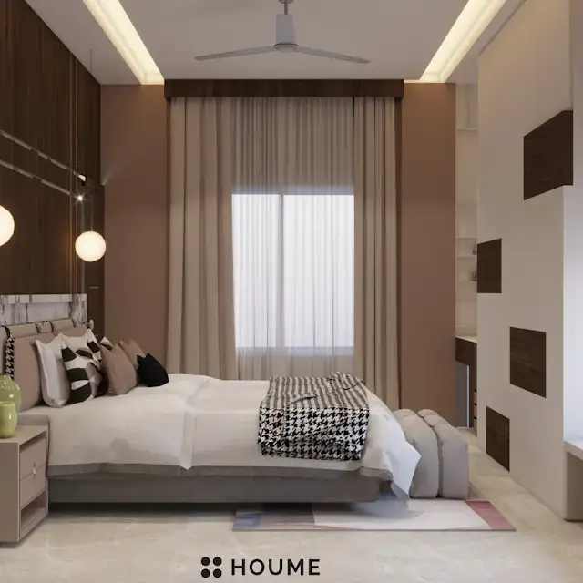 Mid-century modern bedroom interior featuring a platform bed with a walnut wood frame, a woven cream blanket and pillows, and a round wooden nightstand with a drawer. A woven jute rug covers the floor. A ceiling fan with woven blades hangs from the ceiling. The wall behind the bed is painted a light blue color. Text overlay: Houme India - Interior Design Ideas. B