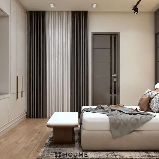 Mid-century modern bedroom featuring a platform bed with a walnut wood frame, a woven cream blanket and pillows, and a round wooden nightstand with a drawer. A woven jute rug covers the floor. A ceiling fan with woven blades hangs from the ceiling. Text overlay: Houme India - Interior Design Ideas. C