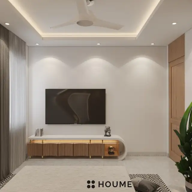 Bedroom interior featuring a mid-century modern platform bed with a walnut wood frame and a woven cream blanket and pillows. A round wooden nightstand with a drawer sits beside the bed. A woven jute rug in a light natural color covers the floor. Text overlay: Houme India - Interior Design Ideas. B