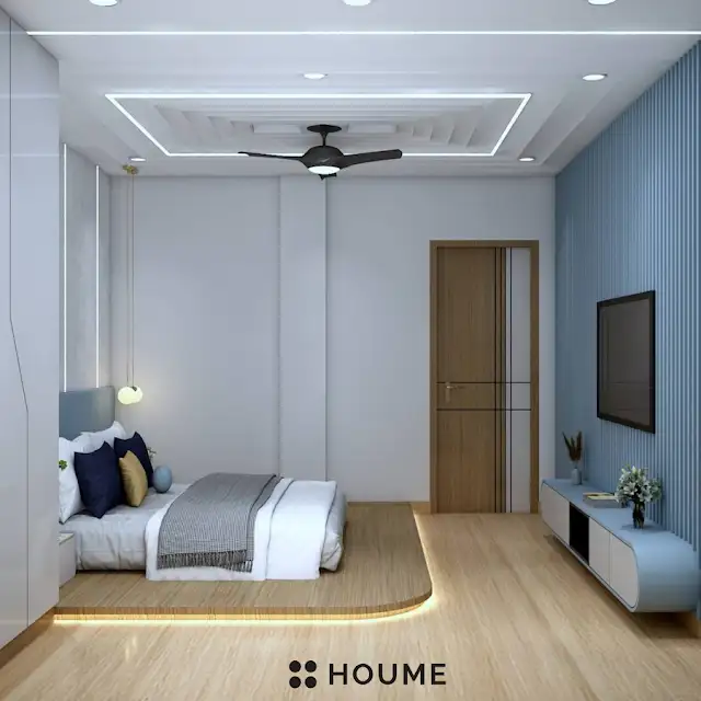 Bedroom featuring a mid-century modern bed with a tufted headboard and tapered wooden legs. A round wooden nightstand with a drawer sits beside the bed. A woven jute rug in a light natural color is on the floor. Text overlay: Houme India - Interior Design Ideas. C
