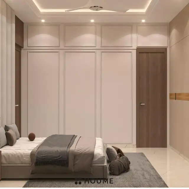 Mid-century modern bedroom interior featuring a platform bed with a walnut wood frame and a woven cream blanket and pillows. A round wooden nightstand with a drawer sits beside the bed. A woven jute rug in a light natural color covers the floor. A ceiling fan with woven blades hangs from the ceiling. Text overlay: Houme India - Interior Design Ideas.B