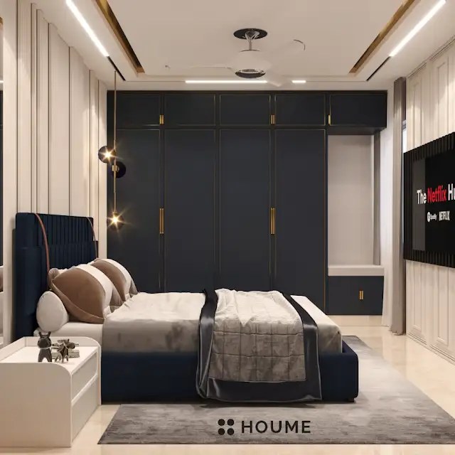 Bedroom featuring a mid-century modern platform bed with a walnut wood frame, a woven cream blanket and pillows. A round wooden nightstand with a drawer sits beside the bed. A woven jute rug in a light natural color covers the floor. A ceiling fan with woven blades hangs from the ceiling. Text overlay: Houme India - Interior Design Ideas. B