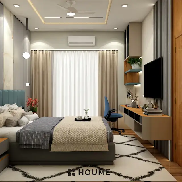 Bedroom interior featuring a mid-century modern platform bed with a walnut wood frame, a woven cream blanket and pillows. A round wooden nightstand with a drawer sits beside the bed. A woven jute rug in a light natural color covers the floor. A ceiling fan with woven blades hangs from the ceiling. Text overlay: Houme India - Interior Design Ideas. B