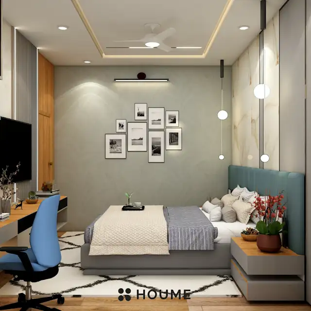 Bedroom interior featuring a mid-century modern platform bed with a walnut wood frame, a woven cream blanket and pillows. A round wooden nightstand with a drawer sits beside the bed. A woven jute rug in a light natural color covers the floor. A ceiling fan with woven blades hangs from the ceiling. Text overlay: Houme India - Interior Design Ideas. C