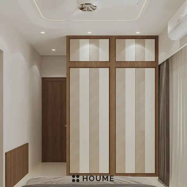 Bedroom featuring a platform bed with a textured white quilt and pillows. A wooden nightstand with a woven storage basket sits beside the bed. A table lamp with a white shade sits on the nightstand. Text overlay: Houme India - Interior Design Ideas. B