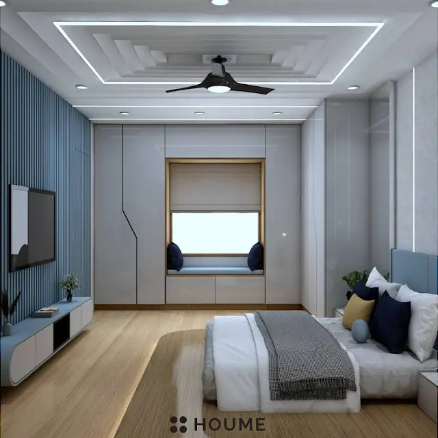 Bedroom featuring a ceiling fan with woven blades hanging from the ceiling. A brown leather couch sits on a woven jute rug in the center of the room. Two brown accent chairs with nailhead trim flank a round wooden coffee table with a black metal base. Text overlay: Houme India - Interior Design Ideas. B