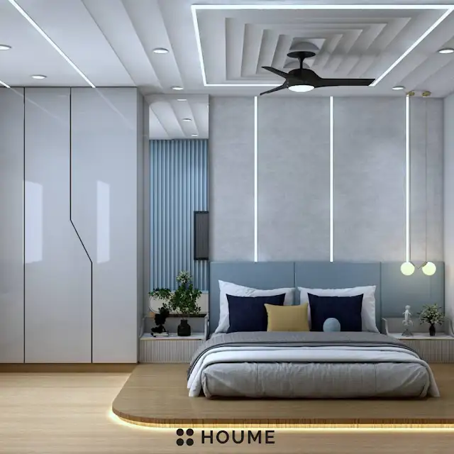 Bedroom featuring a ceiling fan with woven blades hanging from the ceiling. A brown leather couch sits on a woven jute rug in the center of the room. Two brown accent chairs with nailhead trim flank a round wooden coffee table with a black metal base. Text overlay: Houme India - Interior Design Ideas. C