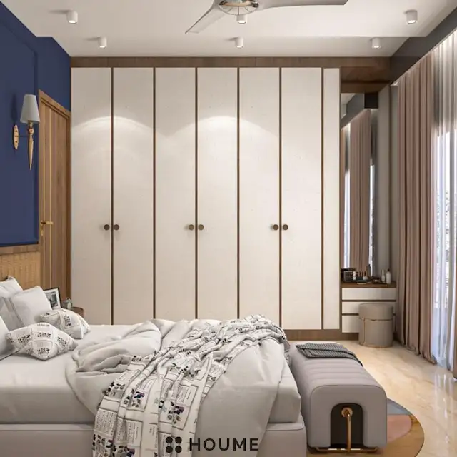 Mid-century modern bedroom interior featuring a platform bed with a walnut wood frame and a woven cream blanket and pillows. A round wooden nightstand with a drawer sits beside the bed. A woven jute rug in a light natural color covers the floor. The wall behind the bed is painted a light blue color. Text overlay: Houme India - Interior Design Ideas. B
