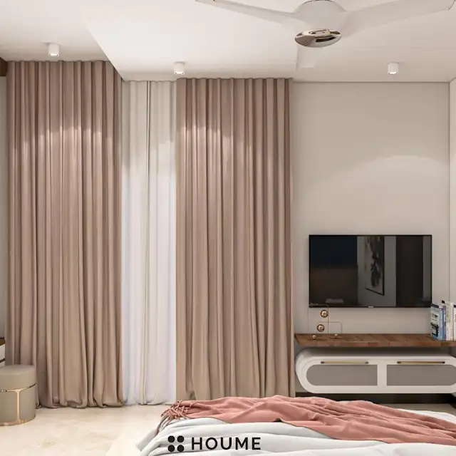 Mid-century modern bedroom interior featuring a platform bed with a walnut wood frame and a woven cream blanket and pillows. A round wooden nightstand with a drawer sits beside the bed. A woven jute rug in a light natural color covers the floor. The wall behind the bed is painted a light blue color. Text overlay: Houme India - Interior Design Ideas. C