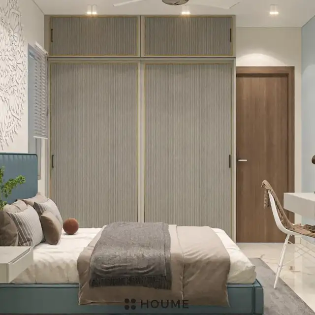Mid-century modern bedroom interior featuring a platform bed with a walnut wood frame and a woven cream blanket and pillows. A round wooden nightstand with a drawer sits beside the bed. A woven jute rug in a light natural color covers the floor. A ceiling fan with woven blades hangs from the ceiling. Text overlay: Houme India - Interior Design Ideas. B
