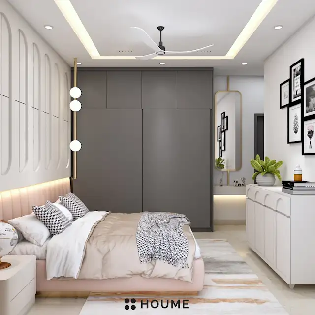Bedroom interior featuring a wooden platform bed with a white quilt and pillows. A vintage world globe sits on a wooden nightstand beside the bed. A ceiling fan with woven blades hangs from the ceiling. Text overlay: Houme India - Interior Design Ideas. C