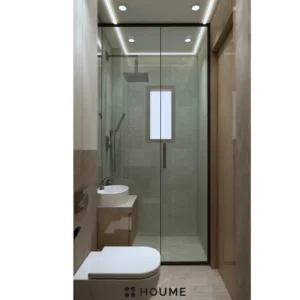 Minimalist bathroom interior featuring a white toilet, pedestal sink with a chrome faucet, and a rectangular mirror with a chrome frame. The walls are a light beige color, and a window with frosted glass on the right wall allows for natural light. Text overlay: Houme India - Interior Design Ideas. C