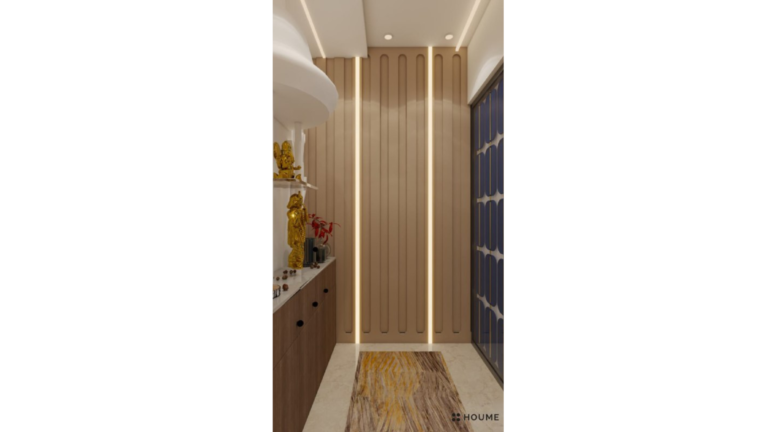 A close-up photo of a modern pooja room design. The pooja unit has a sleek, white marble finish with clean lines and gold hardware accents. The unit features a shelf with a backlit panel displaying Hindu deities. Offerings and prayer items are placed on the shelf. B